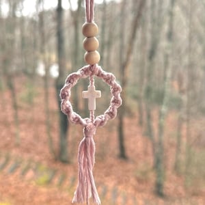 May include: A hanging macrame ornament featuring a dusty pink knotted circular frame with a central white cross charm. The piece is suspended by a matching pink cord adorned with three smooth, round, light-coloured wooden beads and finished with a tassel.