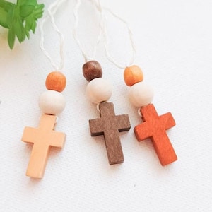 May include: Three wooden cross pendants in shades of orange, brown, and beige, each strung on a white cord with wooden beads. The crosses are displayed against a white background, with a hint of green foliage in the upper left corner.