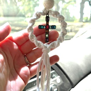 May include: A hanging ornament featuring a white macrame ring with a central cross pendant adorned with multicolored rectangular gemstones. The piece includes a textured round bead at the top and a white cotton tassel hanging from the bottom.