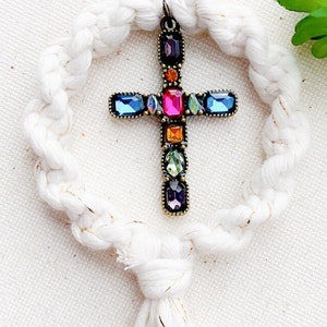 Peut inclure: Un cercle en macramé blanc avec un pompon suspendu en bas. Une croix dorée avec des pierres précieuses colorées pend du centre du cercle.