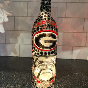 May include: A decorative glass bottle featuring the word "GEORGIA" in red lettering, along with a large "G" logo and a bulldog graphic. The bottle is adorned with black and red embellishments.