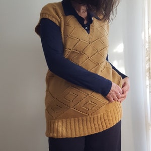 Puede incluir: Chaleco de punto amarillo mostaza con un patrón de diamantes, usado sobre una camisa azul marino con cuello y una camiseta de manga larga. La persona también lleva pantalones negros. El atuendo es informal y cómodo.