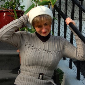 Puede incluir: Una mujer lleva un suéter de punto gris con un gorro de ganchillo blanco y un cinturón marrón. El suéter tiene un delicado patrón de encaje y un cuello en V.