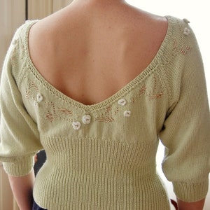 May include: A light green knitted sweater with a low, V-shaped neckline and delicate floral embroidery. The sweater has a fitted bodice and slightly puffed sleeves.