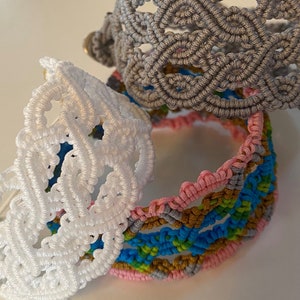 May include: A collection of macrame bracelets in various colors. The bracelets include white, gray, and a multi-colored one with pink, blue, and gold accents. The macrame designs feature intricate woven patterns, showcasing the handmade craftsmanship.