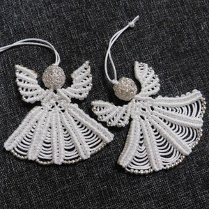 May include: Two handmade white macrame angel ornaments with silver bead accents. Each angel has a spherical head and delicate wings. The wings and skirts are detailed with intricate knotting. A white string is attached for hanging.