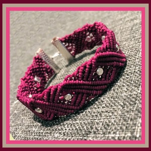 May include: A burgundy macrame bracelet with silver-colored accents. The bracelet features a woven design with small, round silver beads evenly spaced throughout. The clasp is silver-toned. The bracelet is displayed on a gray textured surface.