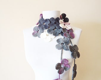May include: A handmade crochet scarf featuring a floral design. The scarf is draped on a mannequin, showcasing a cascade of gray, white, and purple flowers. The flowers are intricately detailed, adding a touch of elegance to the accessory.