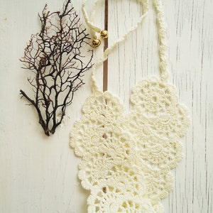 May include: White crocheted necklace with a delicate floral pattern. The necklace is laid out on a white wooden surface with a dried branch to the left.