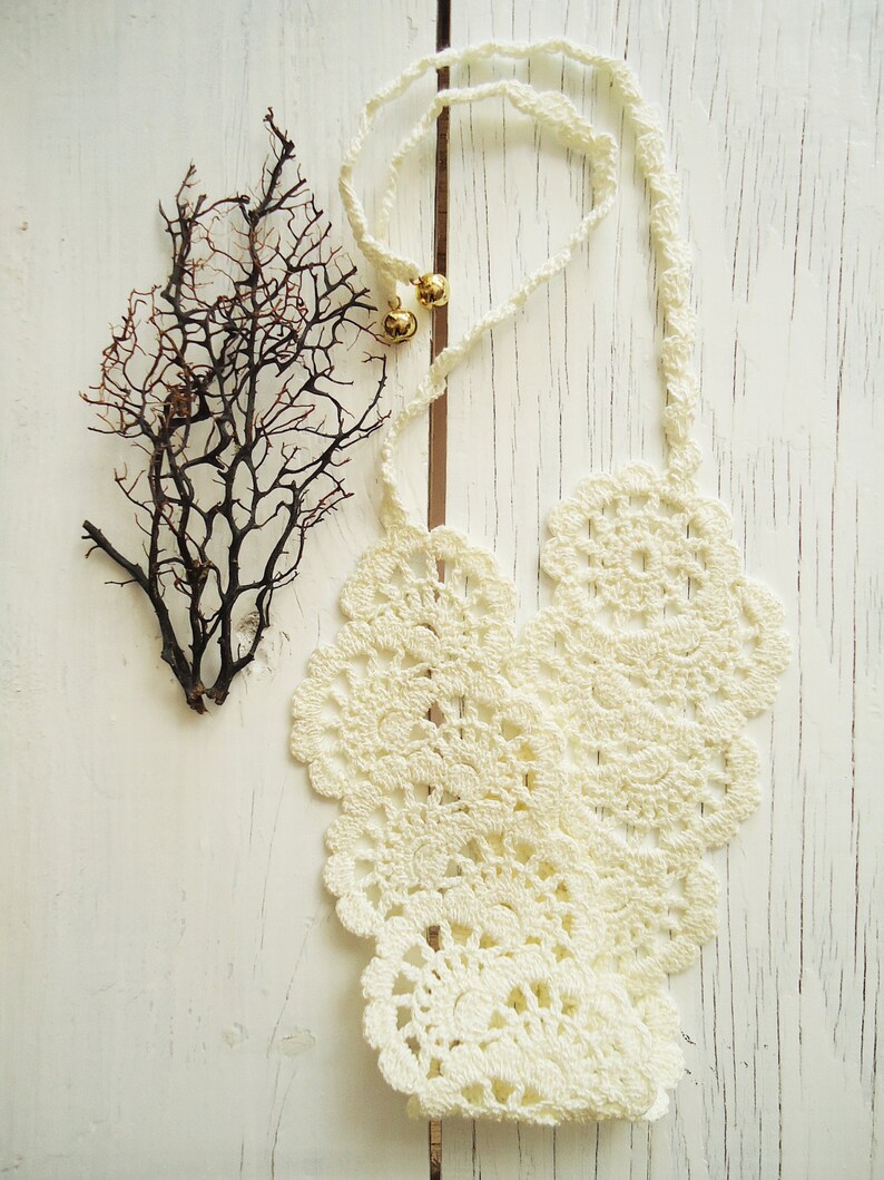 May include: White crocheted necklace with a delicate floral pattern. The necklace is laid out on a white wooden surface with a dried branch to the left.