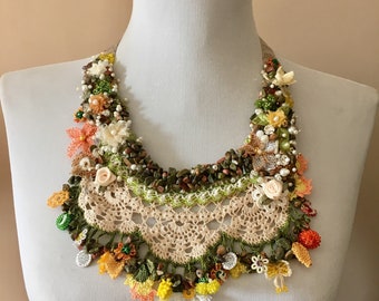 May include: A crocheted necklace with a floral design. The necklace is made with beige, green, orange, and white yarn. It features small flowers, beads, and stones.
