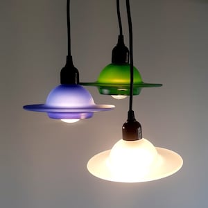 Può includere: Tre luci a sospensione con paralumi in vetro di diversi colori. I paralumi sono verde, blu e bianco. Le luci sono appese a cavi neri.