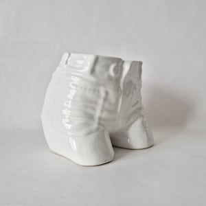 May include: A white ceramic planter shaped like a pair of jeans. The planter has details such as pockets, belt loops, and a button. The legs of the jeans form the base of the planter.