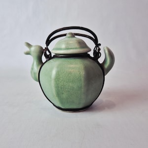 May include: A celadon green ceramic teapot with a black wire handle and trim. The teapot has a lid and a spout shaped like a bird's head. The teapot is approximately 15 cm tall.