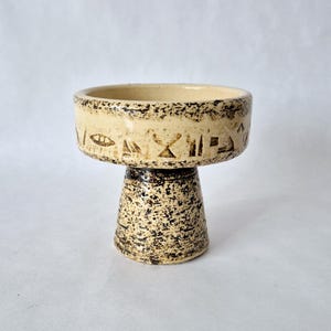 Könnte beinhalten: Beige Keramik-Sockelschale mit strukturierter, gesprenkelter Oberfläche. Die Schale hat eine breite, flache Oberseite mit dunkelbraunen Markierungen und einen Sockel. Der Sockel ist dunkler braun mit einem gesprenkelten Muster.