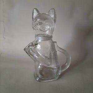 Puede incluir: Una jarra transparente con forma de gato. La jarra presenta una cabeza de gato con orejas puntiagudas y una cara detallada. El cuerpo de la jarra tiene forma de gato, con un asa en el lateral. El vidrio tiene una superficie texturizada.