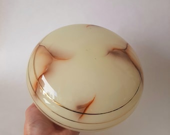 May include: A round, cream-colored glass lampshade with brown and gold marbling. The lampshade has a gold band around the middle and is held by a hand. The background is a plain white.