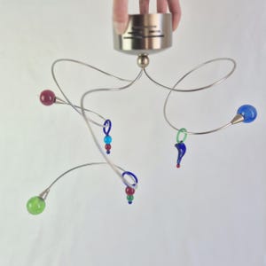 Op de afbeelding: Een moderne plafondlamp met een geborstelde metalen basis en gebogen, zilverkleurige armen. De armen zijn versierd met kleurrijke glazen bollen en decoratieve glaselementen in rood, blauw en groen. De lamp wordt door een hand vastgehouden.