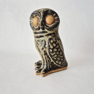 Könnte beinhalten: Eine Keramik-Eulenfigur mit gesprenkelter beige- und dunkelbrauner Glasur. Die Eule hat große, runde, orangebraune Augen und detaillierte Federmuster. Die Figur steht aufrecht und zeigt ihre Füße und ihren strukturierten Körper.