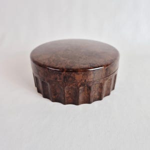 May include: A round, brown jewelry box with a lid. The box has a textured, marbled appearance and a ridged, decorative edge. The box is made of a glossy material and sits on a white surface.