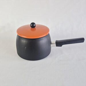 Op de afbeelding: Een zwarte steelpan met een oranje deksel en een zwarte handgreep. Het deksel heeft een zwarte knop. De handgreep is met een zilverkleurig metalen stuk aan de zijkant van de pan bevestigd. De steelpan staat op een witte ondergrond.