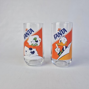 Könnte beinhalten: Zwei transparente Fanta-Gläser mit Disney-Figuren. Ein Glas zeigt Micky und Minnie Maus, das andere Donald Duck. Jedes Glas hat ein orange-weißes Design mit dem Fanta-Logo.