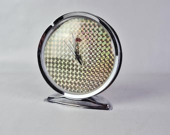 May include: A chrome-finished, round alarm clock with a patterned face. The clock has a silver base and a red accent near the top. The hands are black. The clock is a vintage style.