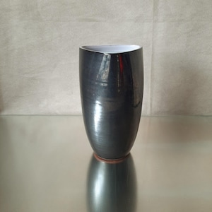 Puede incluir: Un jarrón de cerámica gris oscuro con un acabado brillante. El jarrón tiene una forma ligeramente asimétrica con un interior blanco y una base marrón rojizo. El jarrón se exhibe sobre una superficie reflectante.