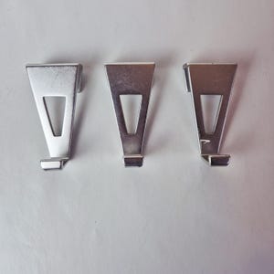 Op de afbeelding: Drie zilverkleurige metalen fotolijsthangers. Elke hanger heeft een driehoekige vorm, een rechthoekige haak aan de onderkant en een V-vormige uitsparing. Ontworpen om fotolijsten op te hangen.