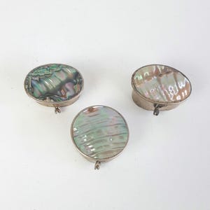 May include: Three small, silver-toned boxes with iridescent abalone shell tops. The boxes are oval and round, with hinged lids and small clasps. The abalone shells display a range of colours, including green, blue, and purple.