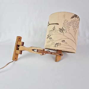 May include: A wooden wall-mounted lamp with a beige lampshade featuring a brown and gray tree branch pattern. The lamp has a natural wood finish and an adjustable arm. The electrical cord is brown.