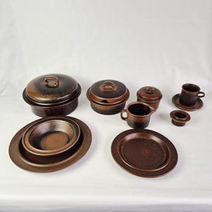 Könnte beinhalten: Eine Sammlung brauner Keramikgeschirr, darunter Servierschalen, Teller, ein Sahnekännchen, eine Tasse mit Untertasse und abgedeckte Schalen. Die Stücke haben ein gesprenkeltes, strukturiertes Aussehen und eine dunkelbraune Farbe.