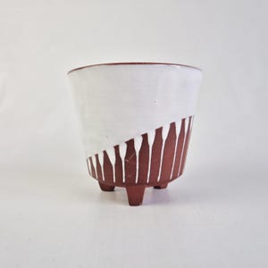 Op de afbeelding: Een kleine keramische plantenbak met drie poten. De bovenste helft is wit, terwijl de onderste helft roodbruin is met witte verticale druppelachtige ontwerpen. De rand en de poten zijn ook roodbruin. Ontworpen voor kleine planten.