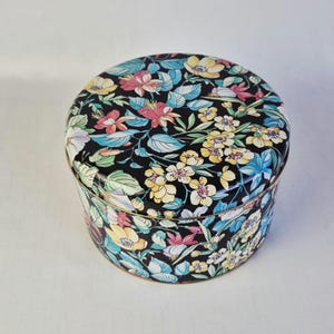 Può includere: Scatola rotonda in latta nera con un motivo floreale. Il coperchio e i lati sono ricoperti di fiori e foglie colorate nei toni del giallo, rosa, blu e verde. La scatola ha un bordo dorato.