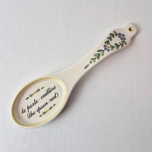 May include: Cream-colored ceramic spoon rest with a floral design and the text "le porte-cuillère (the spoon-rest)". The spoon rest has a yellow border and a hole at the end of the handle for hanging.