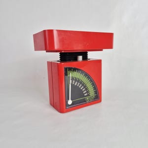 Puede incluir: Una báscula de cocina roja vintage con una base rectangular y un dial curvo verde y blanco. La báscula tiene una plataforma en la parte superior y un dial negro con medidas de hasta 1000 gramos. Una herramienta de cocina clásica.