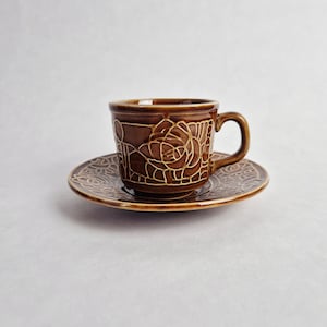 Op de afbeelding: Een bruine keramische kop en schotel set. De kop en schotel hebben een verhoogd bloemmotief in een lichtere bruine kleur. De kop heeft een oor en de schotel is rond. De set staat op een witte achtergrond.