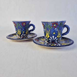Peut inclure: Deux tasses à café et soucoupes en céramique bleues avec des motifs floraux colorés. Les tasses et les soucoupes ont une base bleu foncé avec des motifs floraux jaunes, rouges et blancs. Les tasses ont des anses incurvées et les soucoupes sont rondes.
