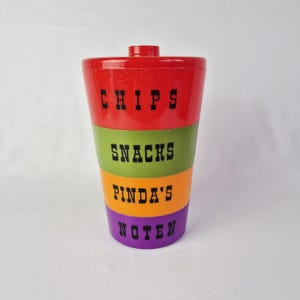 Peut inclure: Un récipient à collations multicolore avec un couvercle et une base rouges, des sections vertes, orange et violettes. Les mots "CHIPS", "SNACKS", "PINDA'S" et "NOTEN" sont imprimés en noir sur chaque section. Le récipient est cylindrique.