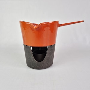 Könnte beinhalten: Orangener Fondue-Topf mit langem Griff und schwarzem Metallständer. Der Topf hat einen Ausguss und steht auf einem dunkelgrauen Metallsockel mit einer kleinen Öffnung für eine Wärmequelle. Ein Klassiker für geschmolzenen Käse oder Schokolade.