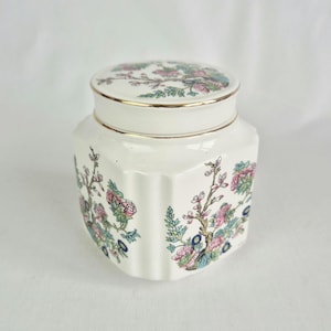 Op de afbeelding: Een witte keramische pot met een deksel, met een bloemmotief in roze, blauw en groen. De pot heeft een gouden rand en een zeshoekige vorm. Het bloemmotief herhaalt zich op het deksel en de zijkanten van de pot.