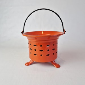 Op de afbeelding: Een oranje metalen kandelaar met een zwarte handgreep. De houder heeft een cilindrisch lichaam met ruitvormige uitsparingen en drie kleine voetjes. De handgreep loopt over de bovenkant.