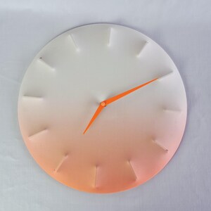 Op de afbeelding: Een ronde wandklok met een wit- naar oranje verloop. De klok heeft oranje wijzers en witte, rechthoekige uurmarkeringen. De diameter van de klok is ongeveer 30 cm.