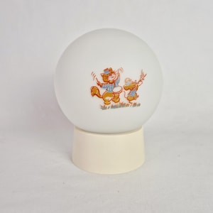 May include: A white, spherical lamp with a cream-colored base. The lamp features a cartoon illustration of a cat playing a drum and a mouse playing a flute. The illustration is in shades of orange, blue, and brown.