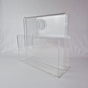 May include: A clear acrylic magazine rack with a rectangular handle and a circular opening. The rack has three vertical dividers and a rectangular base. The design is modern and minimalist.