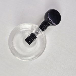 May include: A clear glass bottle with a black stopper. The stopper has a round top and a threaded stem that screws into the bottle. The bottle is empty and sits on a white surface.