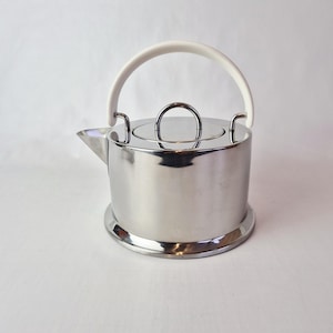 Op de afbeelding: Een roestvrijstalen theepot met een witte handgreep en een kleine, ronde dekselgreep. De theepot heeft een tuit en staat op een cirkelvormige basis. Het ontwerp is minimalistisch en modern.