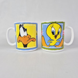 May include: Two white ceramic mugs featuring cartoon characters. One mug displays Daffy Duck on a yellow background, the other shows Tweety Bird on a blue background. Both mugs have white handles and blue and green borders.