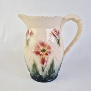 Puede incluir: Jarra de cerámica color crema con diseño floral. La jarra tiene asa y pico vertedor, con flores en relieve en tonos rosa, rojo y verde. La base es verde oscuro y la jarra tiene una estética vintage.