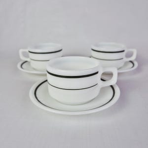 Op de afbeelding: Set van drie witte keramische kopjes en schotels. Elk kopje en schotel heeft een zwart streepontwerp. De kopjes zijn afgerond met een handvat. De schotels zijn rond met een zwarte rand.